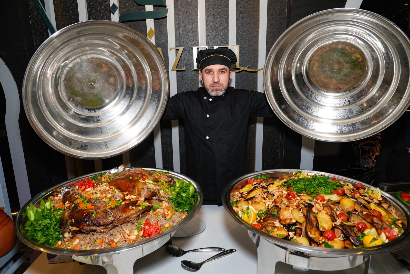 A Jazeel chef behind two large platters — roast lamb on spiced rice, and vegetables with potato and aubergine